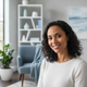 Headshot of a smiling woman in her late 20s to early 30s, with a blurred background of a modern living space.